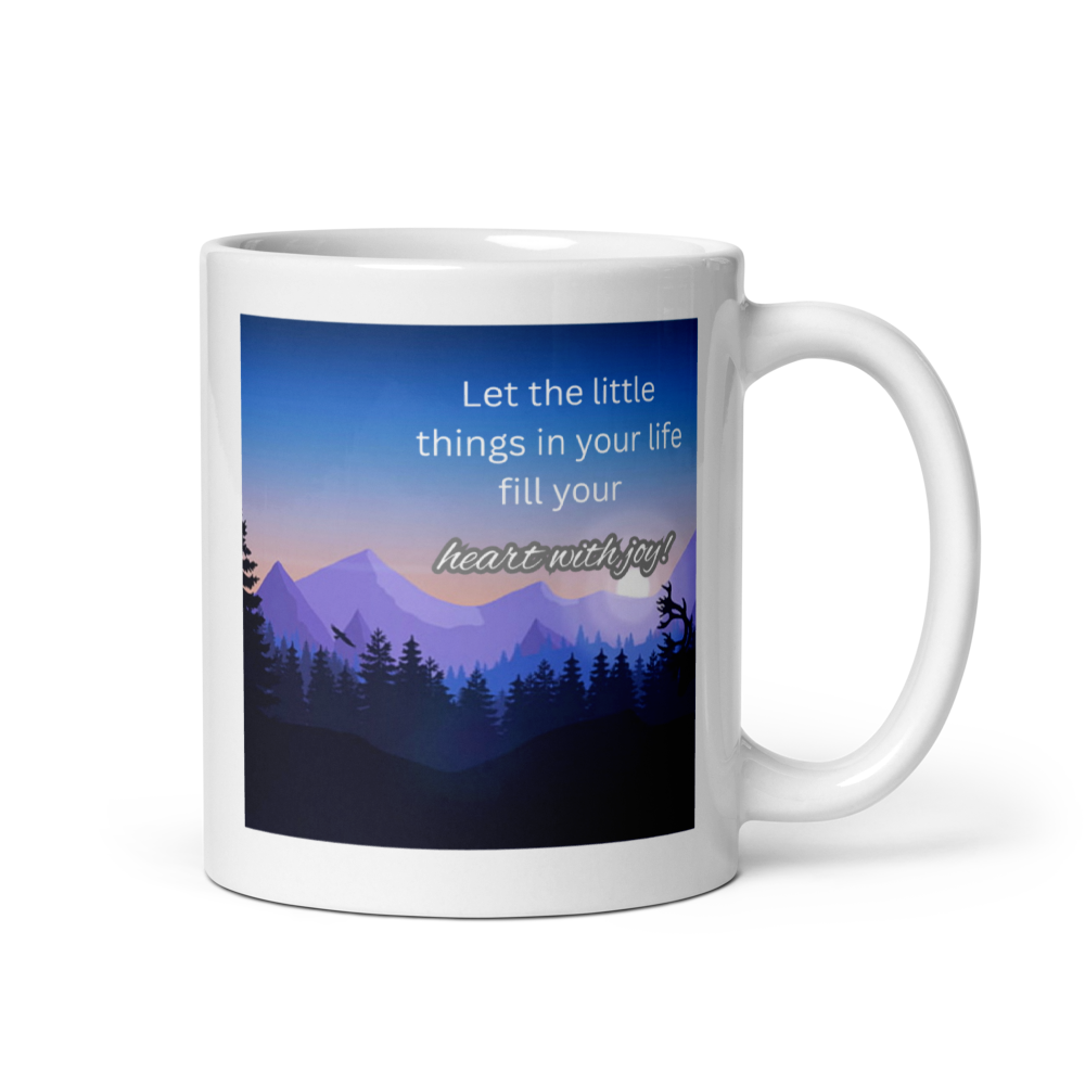 A white ceramic mug with a printed graphic of a mountain landscape at dusk or dawn. The graphic features a blue and purple sky, silhouettes of pine trees, and the text 'Let the little things in your life fill your heart with joy!'.
