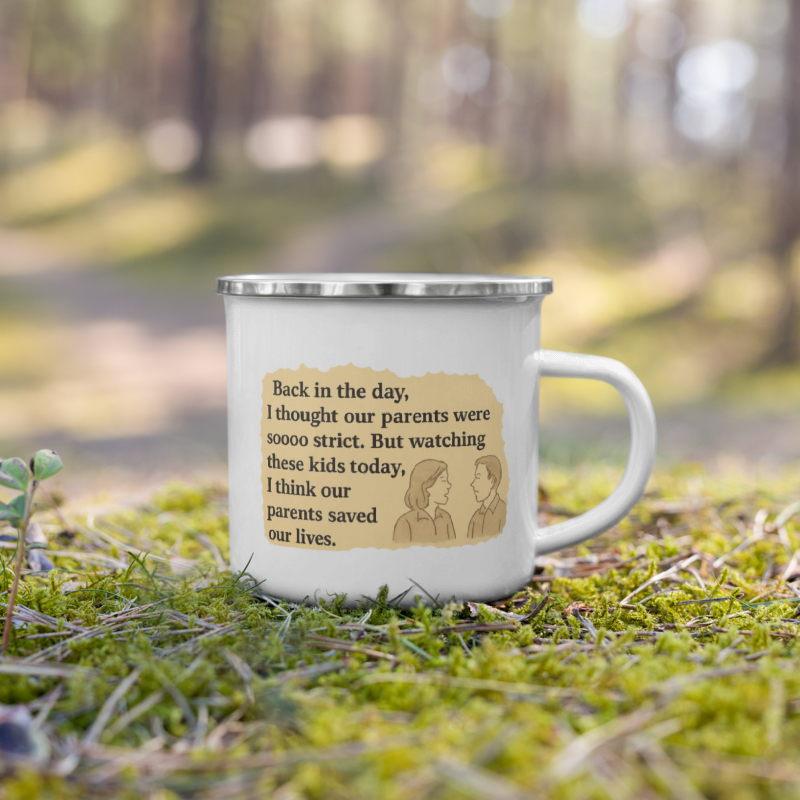 A white enamel camping mug with a silver rim sits on green moss in a blurred forest setting. The mug features a printed beige graphic with a handwritten note design containing a humorous parent-child quote.