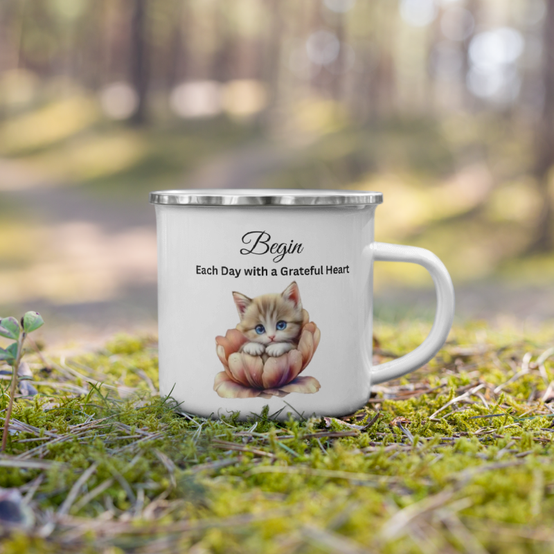 A white enamel camping mug with a silver rim sits on green moss in an outdoor forest setting. The mug features a printed illustration of a cute orange kitten sitting in a flower, with the text 'Begin Each Day with a Grateful Heart' printed above it.