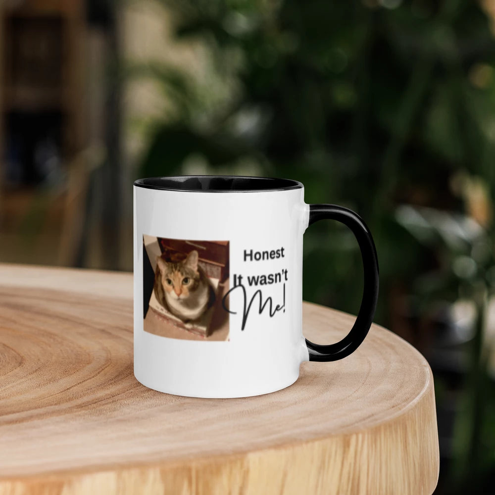 A white ceramic coffee mug with a black handle and interior, featuring a printed photo of a cat and the text 'Honest It wasn't Me!' on a light wood surface with a blurred green background.
