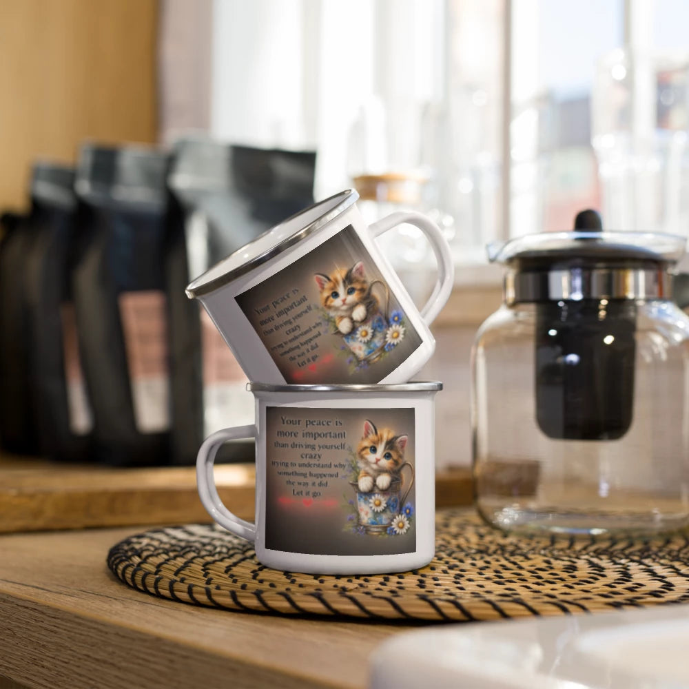 Two stacked white enamel-style mugs with a printed kitten illustration and quote on the front, placed on a woven placemat on a wooden table in a kitchen setting.