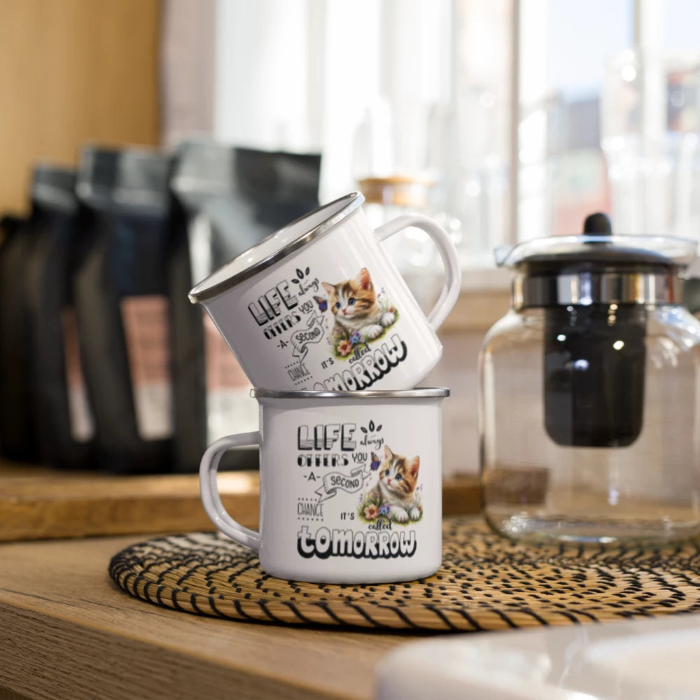 Two stacked white enamel-style mugs with black text reading 'LIFE ONLY TODAY' and a kitten illustration, placed on a woven placemat on a wooden table with coffee bags and a glass teapot in a kitchen setting.