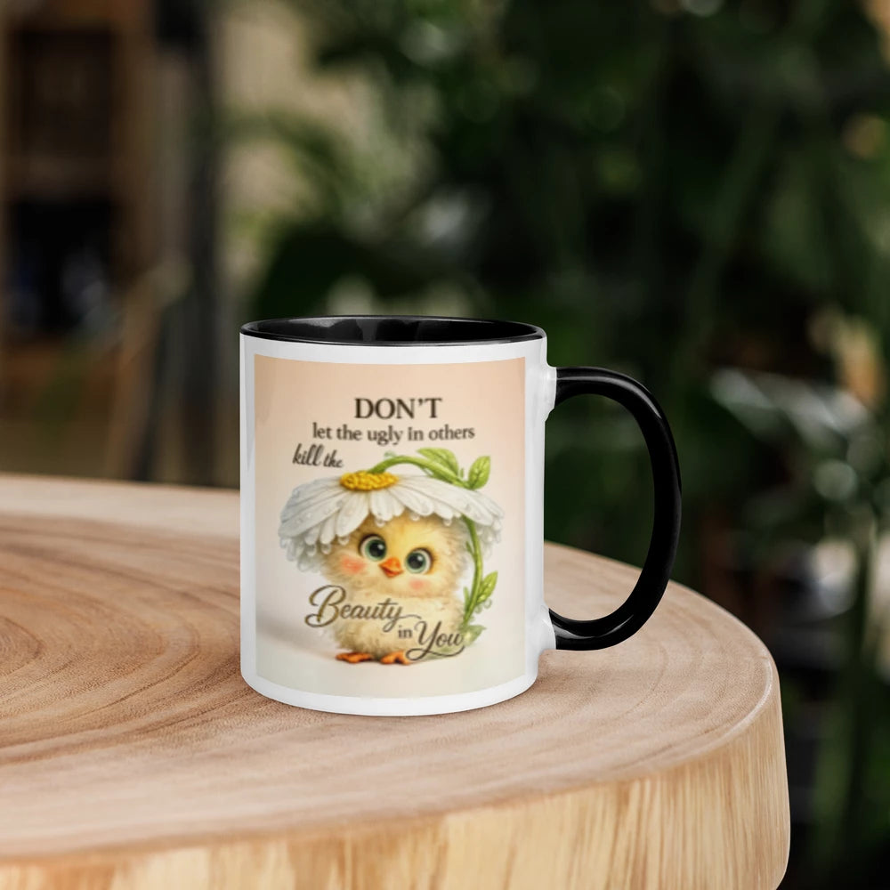 A white ceramic mug with a black handle and interior sits on a round wooden table. The mug features a printed illustration of a cute yellow chick wearing a daisy flower, with the text 'DON'T let the ugly in others kill the Beauty in You'.