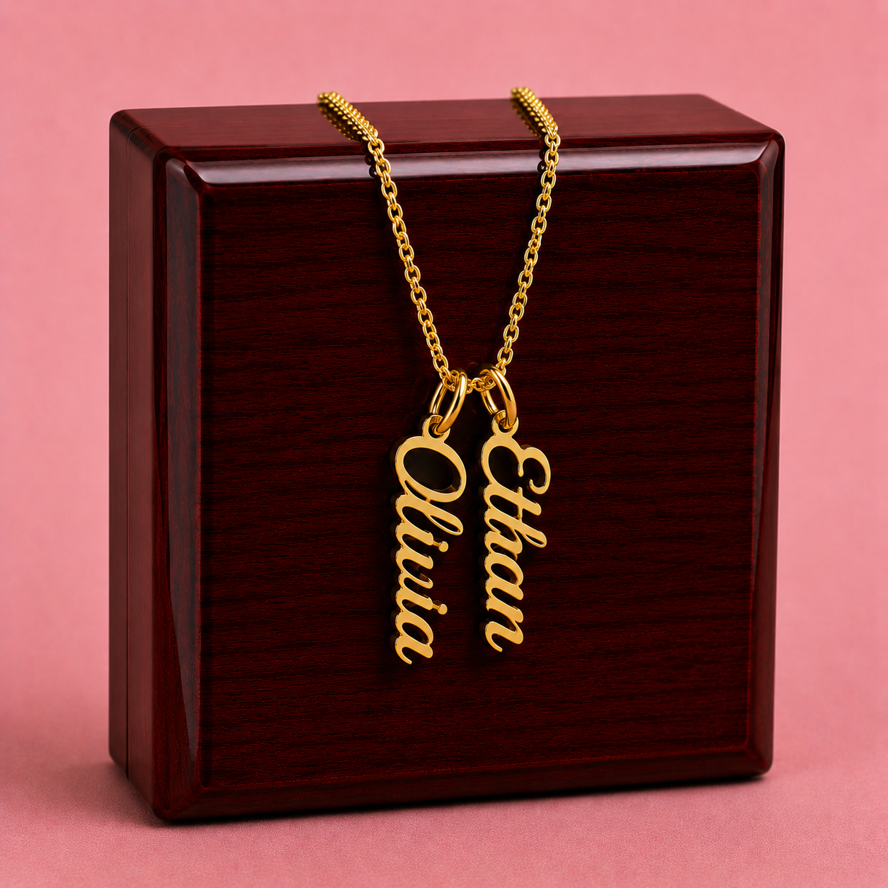 Gold necklace with 'Olivia' and 'Ethan' charms on a wooden box with a pink background