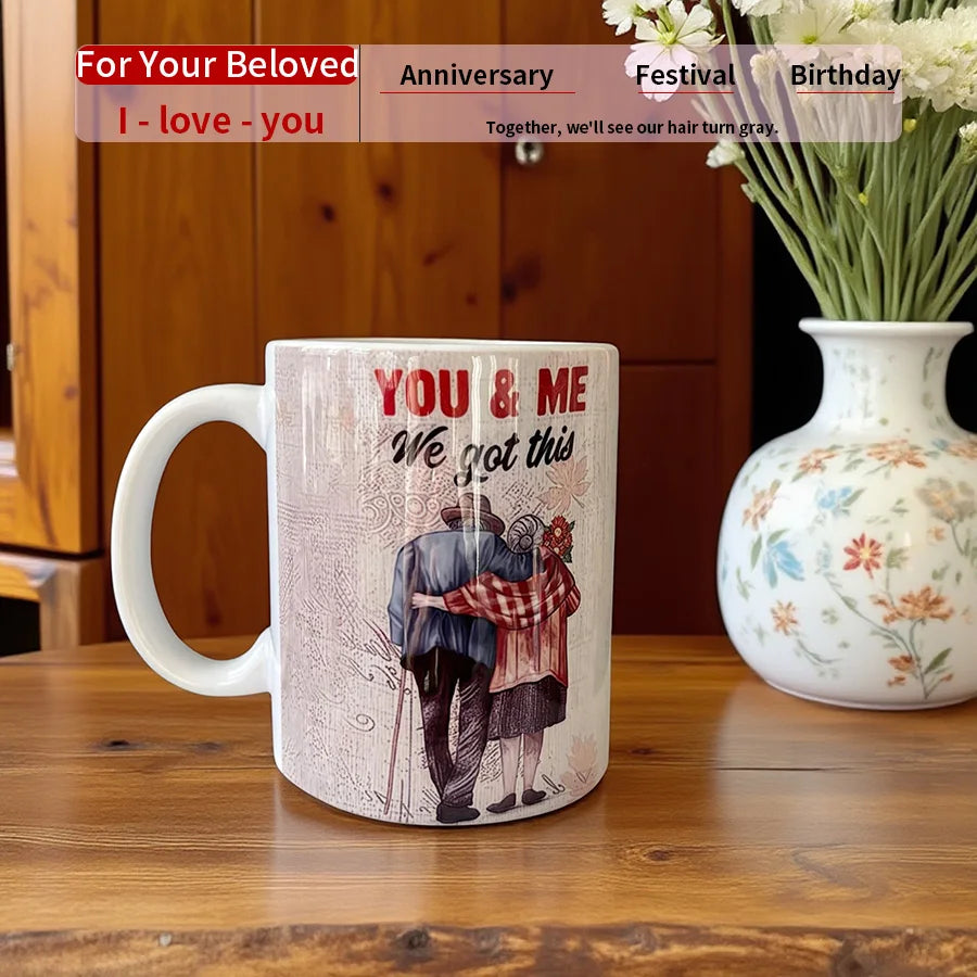 Mug with 'You & Me, We got this' text and a couple illustration on a wooden table with a vase of flowers.