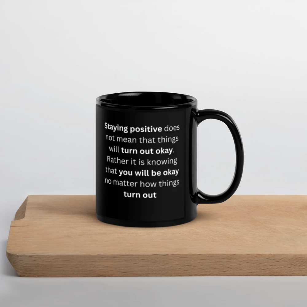 A black ceramic mug with white printed text sits on a light wood board against a clean white background.