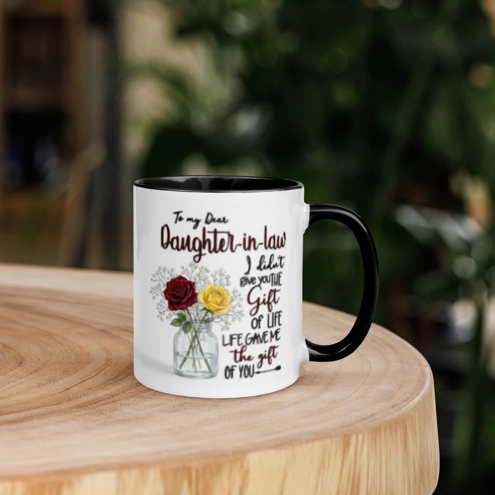 A white ceramic mug with a black handle and interior, printed with a sentimental message to a daughter-in-law and an illustration of red and yellow roses in a vase, displayed on a wooden table with a blurred green garden background.